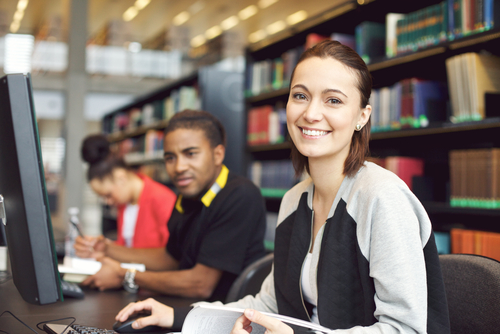 Beautiful young caucasian student sitting at table with computer looking at camera smiling. Young university students researching information on computer for their studies.