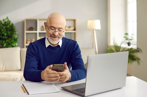 Senior man using mobile phone. Happy retired man in glasses sitting at desk with laptop and cellphone, paying bills online, reading news, sending emails, making banking transaction, calling his