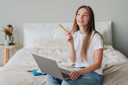 Creative web designer works at home using laptop sitting on bed holds pen looks up gets idea. Pretty Italian student girl remote learning. Distance education. Blonde caucasian woman planning business.
