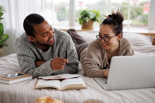 positive multiracial couple in sweaters talking during online courses near devices and books on bed at home