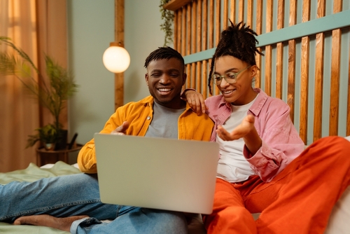 Portrait of smiling happy couple, African American young woman and man using laptop sitting on comfortable bed, watching video, making online ordering working together at home. Technology concept