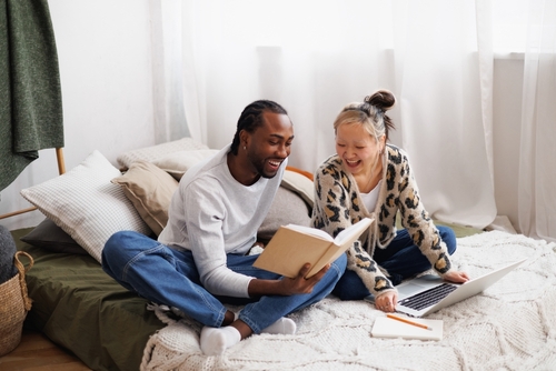 Excited young multiethnic couple reading book near laptop on bed at home