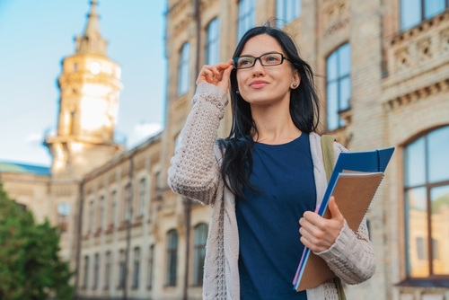 University college female student woman in eyeglasses casual clothes smiling with with books notebooks and bag standing outside near on campus building outdoor and looking away. Education concept