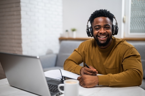 Portrait of young cheerful man with wireless headphones attending online course via laptop and writing notes while sitting at the desk in his home.