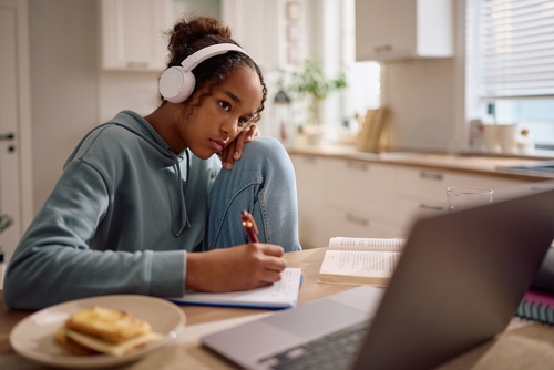Bored African American student taking notes in her notebook while studying over laptop at home.