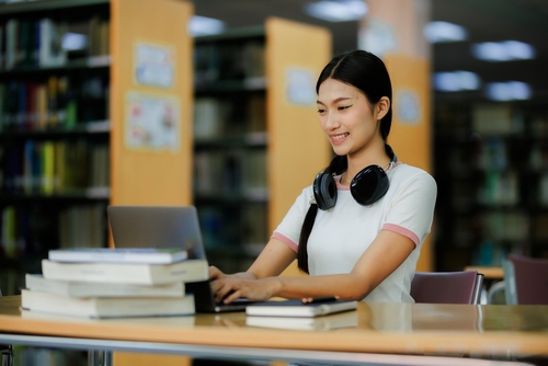 A dedicated female student spends her free time calmly researching in the library, wearing headphones and enjoying music while focusing on her studies.